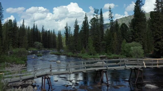 Drone flying along wooden bridge over clear mountain lake in Rahmanovskie Springs, Altai, East Kazakhstan. Scenic pine forest, calm water and alpine landscape in remote wilderness.