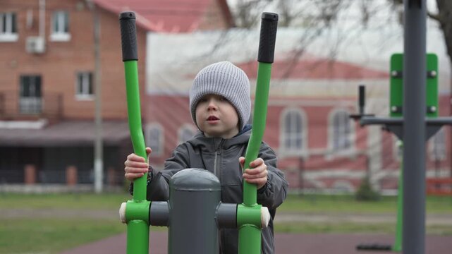 Cute little boy in a hat and jacket training on a public elliptical machine at an outdoor gym, actively engaging in physical fitness and enjoying a healthy lifestyle in a city park setting