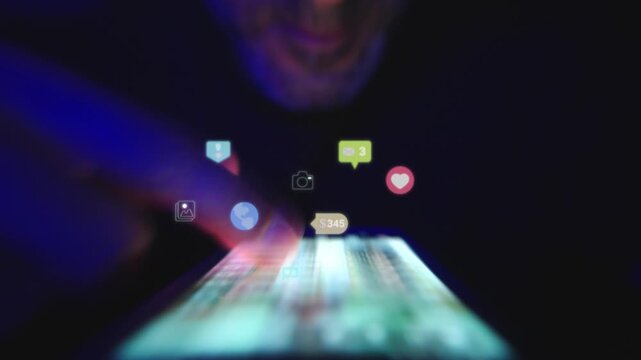 Close up of hands scrolling through a social media feed at night with glowing reaction icons above the screen