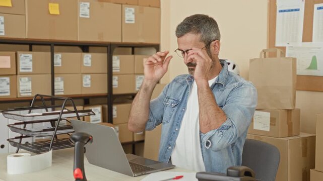 Man covers face with hands at laptop amid stacked cardboard boxes, tape and scanner in building; stress business strain.
