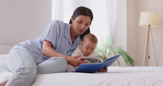 Young 30s attractive mother reading book or showing pictures to her cute little baby while lying together on bed in cozy bedroom at home. Early kid education, connection and development concept 