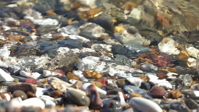 Crystal clear water from a gentle wave flows and ripples over colorful, smooth pebbles on a sunny day, creating a beautiful and tranquil natural background scene with sparkling sunlight reflections