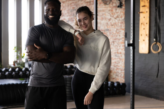 Diverse couple posing and smiling in gym wearing activewear beside dumbbells, squat rack and rings