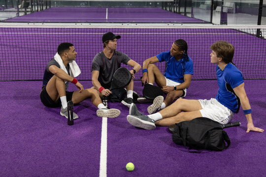 Diverse male teammates sitting on purple padel court near net, in blue shirts and holding paddles