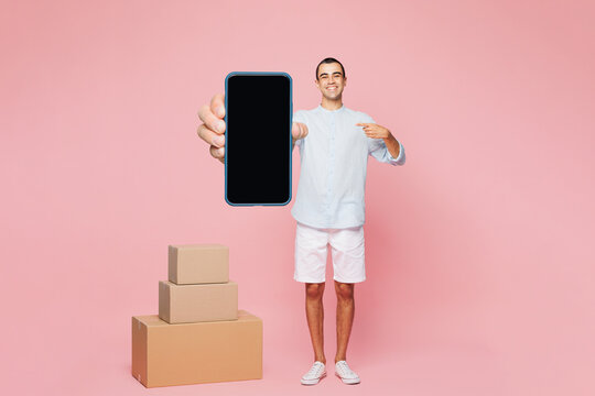 Full body young middle eastern man wear casual blue shirt hold in hands stack cardboard blank boxes delivery use mobile cell phone with blank screen workspace area isolated on plain pink background