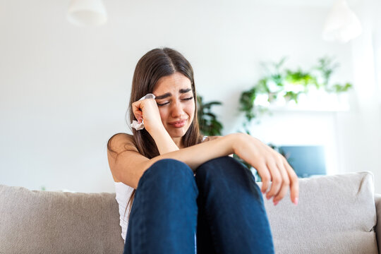 Sad young woman crying and wiping tears with tissue while sitting on sofa. Depression, Mental health or Heartbreak concept. Unhappy girl feeling lonely, stressed and suffering from emotional pain.