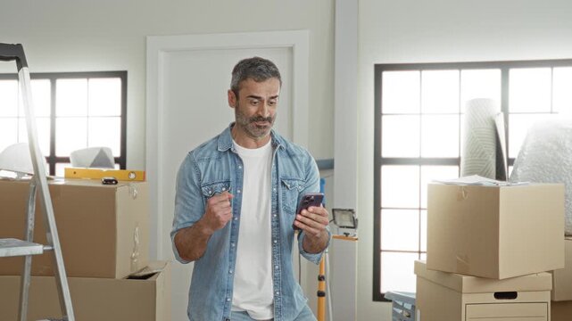 Middle age man pumps fist and smiles while holding smartphone among stacked moving boxes inside a bright building; joy new home.