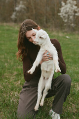 Fototapeta premium A woman gently holds a young goat in a grassy meadow, capturing a tender lifestyle moment of affection and care between human and animal in a natural rural setting.