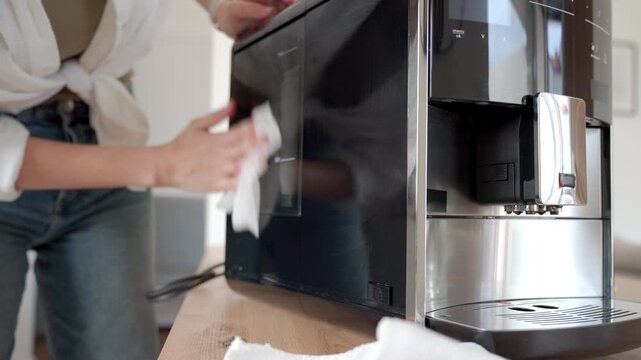 Woman wipes side panel of black coffee machine with cloth on kitchen counter. Home appliance cleaning hygiene maintenance and coffee equipment care