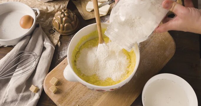 Woman hands pouring white flour from glass jar into bowl with yellow cake batter. Baker preparing homemade sweet dough on rustic wooden board with eggs and whisk. Culinary process of adding dry