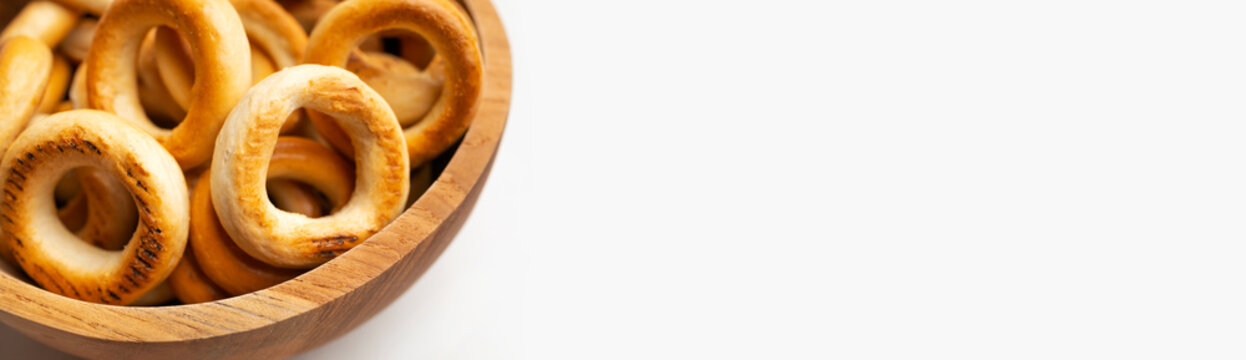 Close-up small bagels in wooden bowl on white background, top view. Freshly baked round mini buns, dry cookies for tea, delicious flour-based pastries. Long banner with space for text