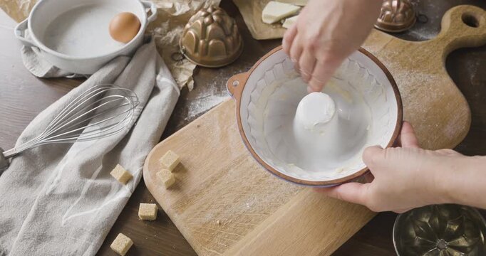 Woman hands greasing white ceramic baking pan with butter on rustic wooden board. Baker preparing cake mold for homemade sweet dessert with flour eggs and whisk in kitchen. Culinary process of coating