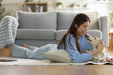 Fototapeta premium Happy asian woman play with pet dog on room floor. Smiling young adult female enjoy comfortable home lifestyle with cute animal companion relaxing indoors bringing deep joy