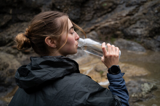 Side view portrait of young Caucasian woman traveler drinking water outdoors while hiking in the mountains.
