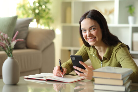 Happy young woman writing in agenda holding phone looks at you
