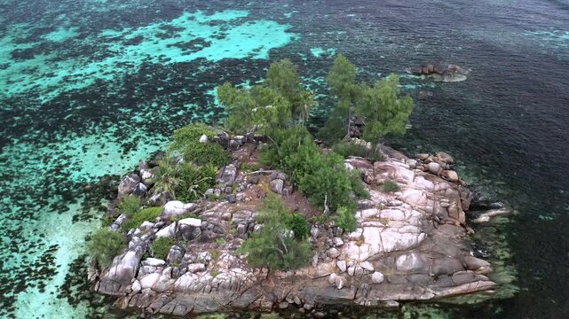Aerial medium-height drone shot of a small rocky granite islet covered with trees, surrounded entirely by turquoise shallow reef and deeper ocean in the Seychelles.