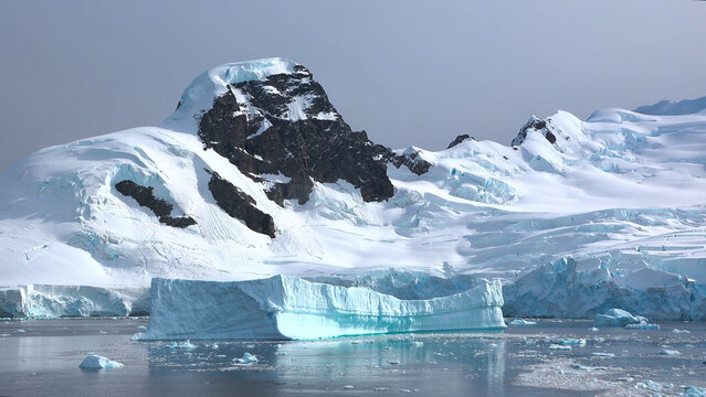 A flat-topped, brilliant blue iceberg floats serenely in the foreground, anchored within a vast field of smaller ice fragments in the frigid Antarctic waters.

Majestic snow-draped peaks and dark, roc