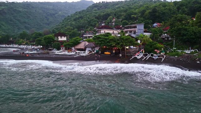 Drone aerial of the traditional Amed fishing village shoreline with white-crested waves washing onto a black volcanic sand beach lined with jukung outrigger boats beneath lush green hillsides. Bali.