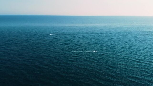 High aerial drone shot over the open Mediterranean Sea. Two small boats travel in opposite directions, their faint white wakes tracing parallel lines across the deep blue water under a soft pastel sky