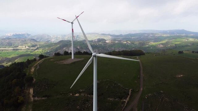 Slow orbital drone shot around two wind turbines at the Parco Eolico di Mineo, Sicily. Rolling green hills, rocky outcrops and snow-dusted mountains visible across the Sicilian interior landscape.