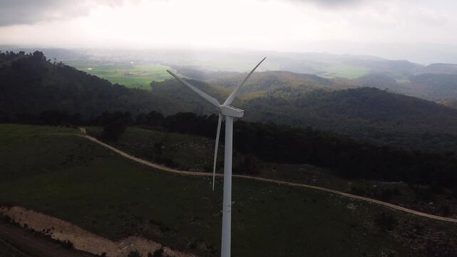 Parallax drone pullback shot revealing a wind turbine at the Parco Eolico di Mineo, Sicily. As the drone reverses, misty forested hills and green farmland gradually unfold behind the rotating blades.