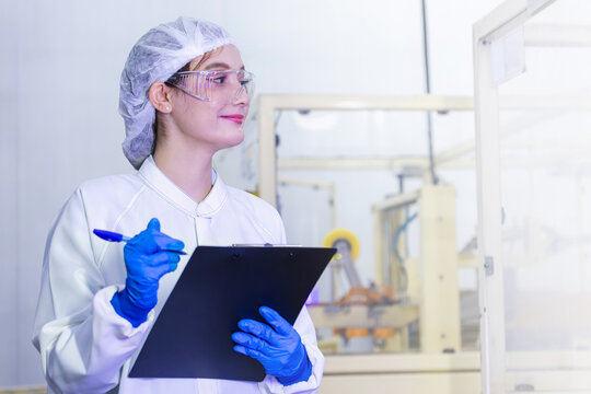 Smiling female quality control inspector in white lab coat holding clipboard in factory, Professional woman engineer in protective gear taking notes during industrial production audit