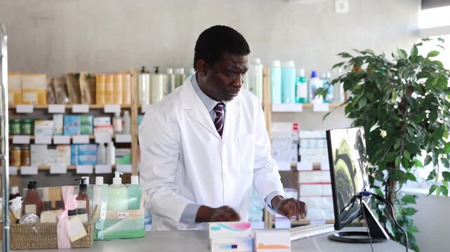 African man pharmacist uses computer at workplace. Staff reads instructions on computer, interprets medical prescription and makes list of drugs to sell 