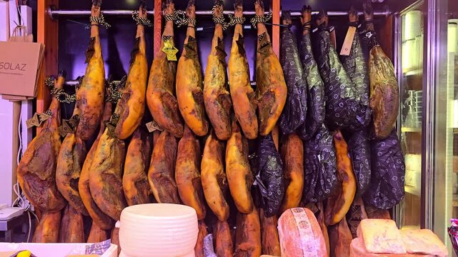 Close up view of Spanish jamon legs hanging on hooks with cheese and sliced ham on a market stall, showing rich texture, traditional curing and authentic local food culture in a vibrant market scene.