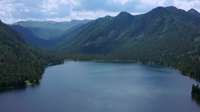 Cinematic drone flyover above Rakhmanovskoye Lake framed by forested Altai mountains in East Kazakhstan. Calm water, deep valley and dramatic ridgelines form pristine alpine wilderness scenery.