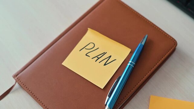 A hand places a sticky note labeled PLAN on a brown notebook. A blue pen lies beside it, signaling the start of an organized approach to tasks in a workspace