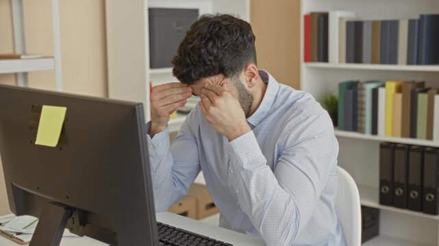Man reading papers and rubbing eyes at desk with computer monitor, sticky note and bookshelves in office; fatigue concentration paperwork deadline.