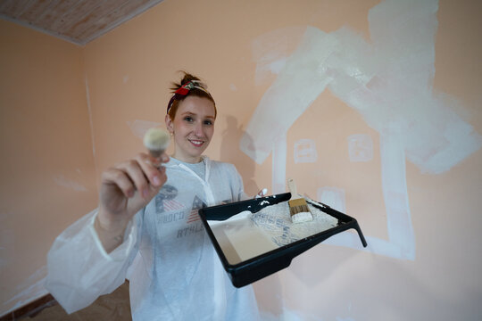 Smiling painter holding brush indoors by wall with painted house