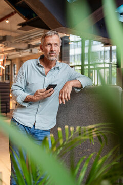 Thoughtful man with smartphone in modern office leaning on partition