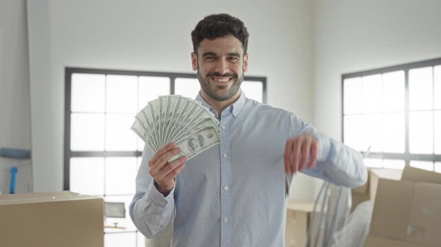 Man holding a fan of cash and dangling keys with right hand raised in building, smiling and posing; financial success.