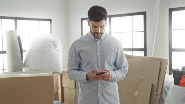 Young hispanic man holding smartphone among stacked moving boxes, bubble wrap and a rolled rug in an empty apartment building, checking messages on his phone; moving planning optimism.