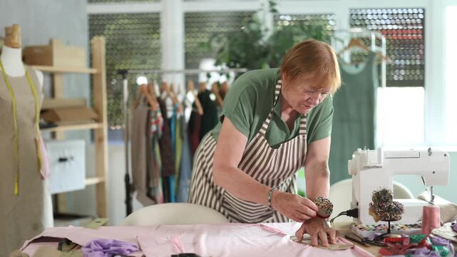  Elderly female seamstress cutting fabric with scissors in sewing workshop