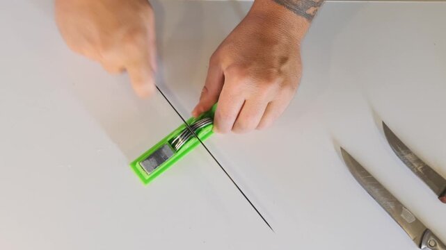 male hands sharpening kitchen knife on manual sharpener top view on table, restoring blade edge for cooking, maintenance process, kitchen routine, tool care and food preparation readiness