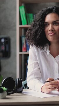 A young woman with curly hair records herself on a smartphone while writing notes at a desk with a laptop and books