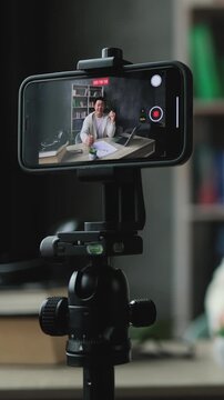 A man is recording himself on a smartphone mounted on a tripod, possibly for a vlog or online course, with a laptop and bookshelf in the background