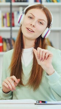 Cheerful teenage student girl in headphones singing song while doing homework in library. Joyful female student distracted from studying