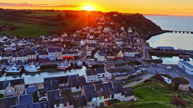 Aerial of a picturesque Normandy fishing village and harbor glowing under a blazing golden sunset over the sea. Normandy, France, Port-en-Bessin.