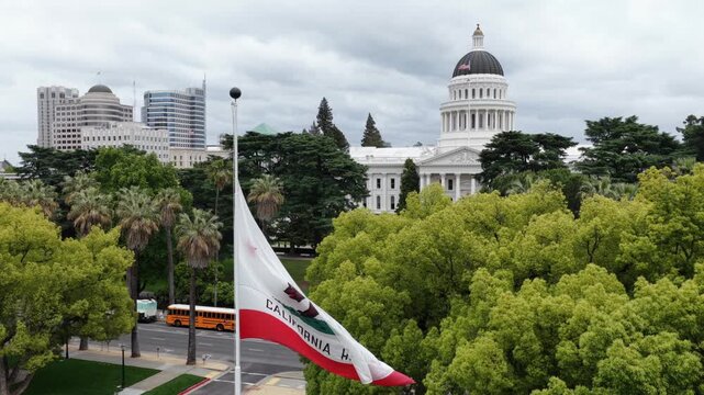 Aerial view of California State Capitol in Sacramento with waving CA state flag, green spring trees and downtown skyline under cloudy skies, showcasing patriotic USA landmark.