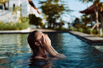 Fototapeta premium Woman in a pool, hands over her face, enjoying relaxation and self care by clear water and lush greenery, warm lighting, tranquil lifestyle moment in a sunny outdoor setting with calm reflections.