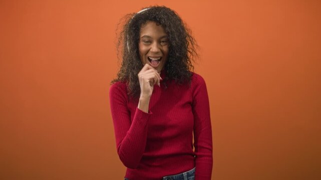 Teen woman with curly hair in red sweater and denim jeans, hand near mouth and open smile, wearing white headband headphones in a studio orange backdrop; joyful surprise.