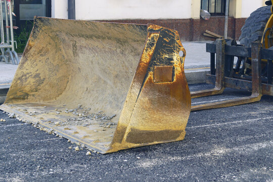 Front-loader bucket resting on dark fresh asphalt beside residential building entrance. Highlights municipal resurfacing encroachment, contractor machinery staging, and active roadway works.