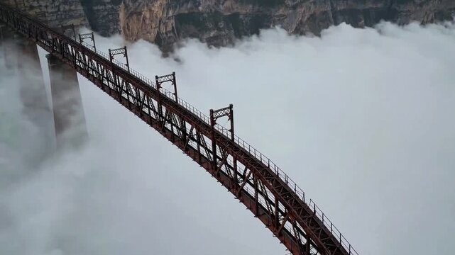 High metal bridge hanging over thick white clouds and deep fog, construction connecting mountain peaks creates sense of adventure, industrial structure rises above morning mist in wild landscape