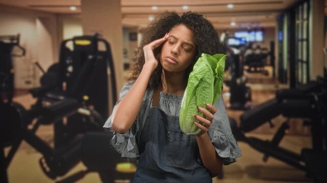 Woman in apron holding romaine lettuce and hand to temple for headache inside a gym building, eyes closed and pained; fatigue health concern irony.