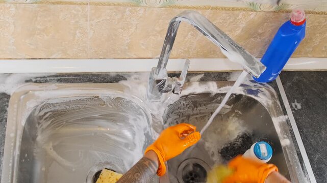 Top view of man wearing gloves scrubbing kitchen sink and faucet with sponge and cloth, removing dirt and soap residue under running water, household cleaning routine in modern home.