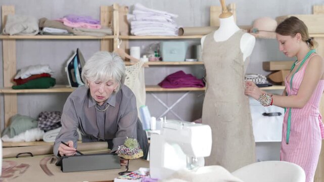 Positive mature tailoress drawing dress model in tablet while other working with mannequin in background. High quality 4k footage