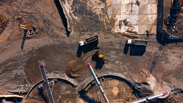 Aerial view of trucks loading at industrial aggregate plant. Industrial scene showing front loader and trucks moving crushed stone near conveyor belts at a mining facility.
